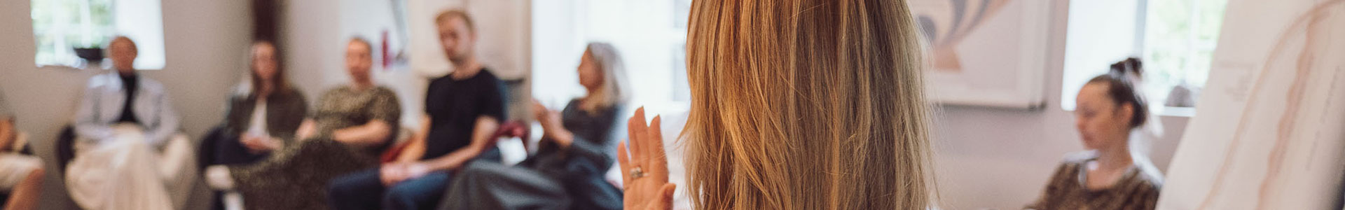 Undervisningssituationen på psykoterapeutisk uddannelse Tobias‑Skolen, hvor en person sidder forrest og henvender sig til en cirkel af deltagere i et lyst undervisningslokale med terapeutiske materialer og visuelle plancher.