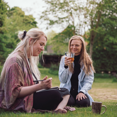 To personer sidder i græsset og deler en rolig stund med notesbog og drikke i grønne omgivelser – illustrerer fællesskab, nærvær og personlig udvikling som en del af Tobias‑Skolens psykoterapeutuddannelse og efteruddannelser.